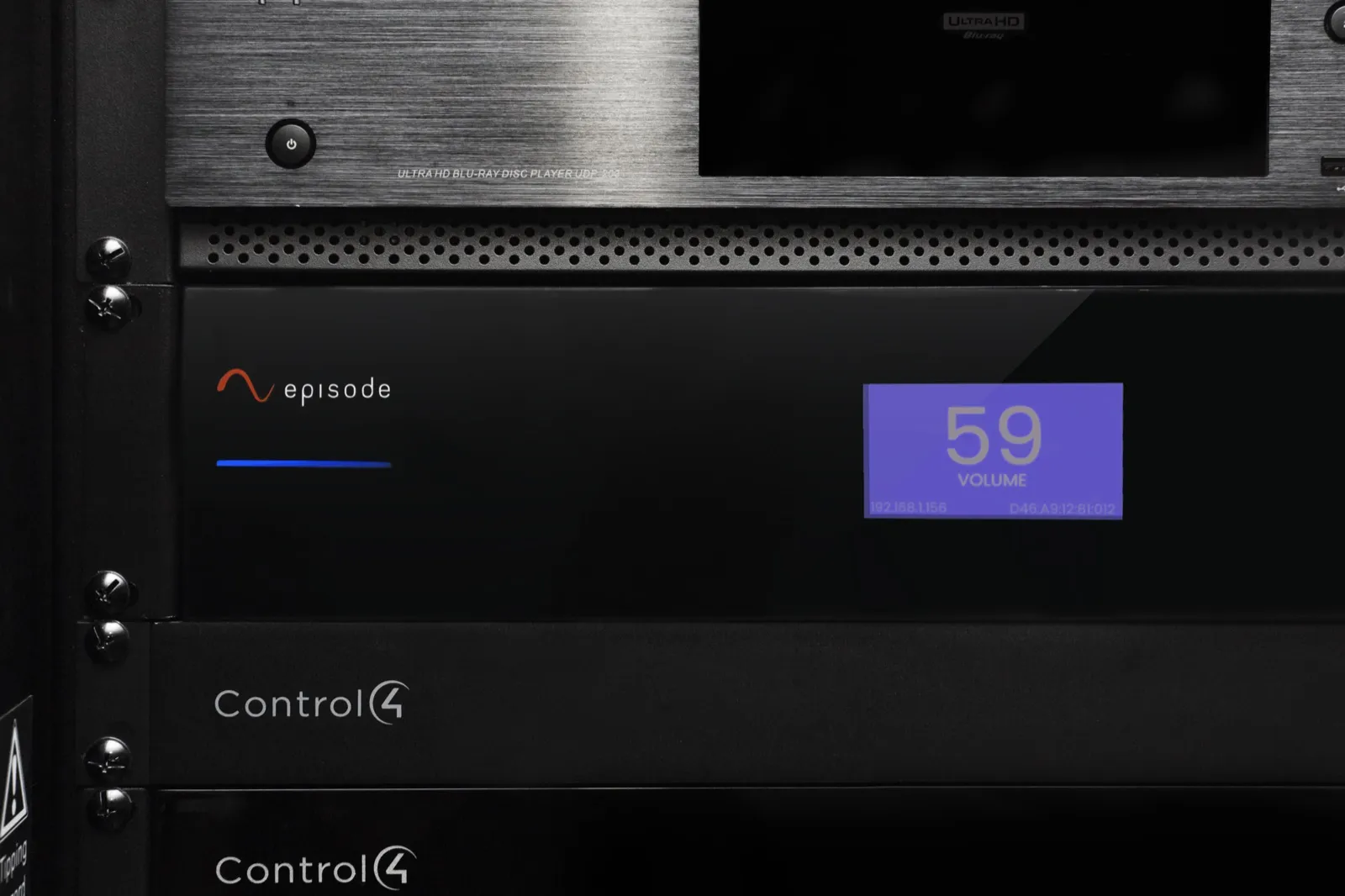 An Episode amplifier in an equipment rack, its front screen showing volume 59, above two Control4 units
